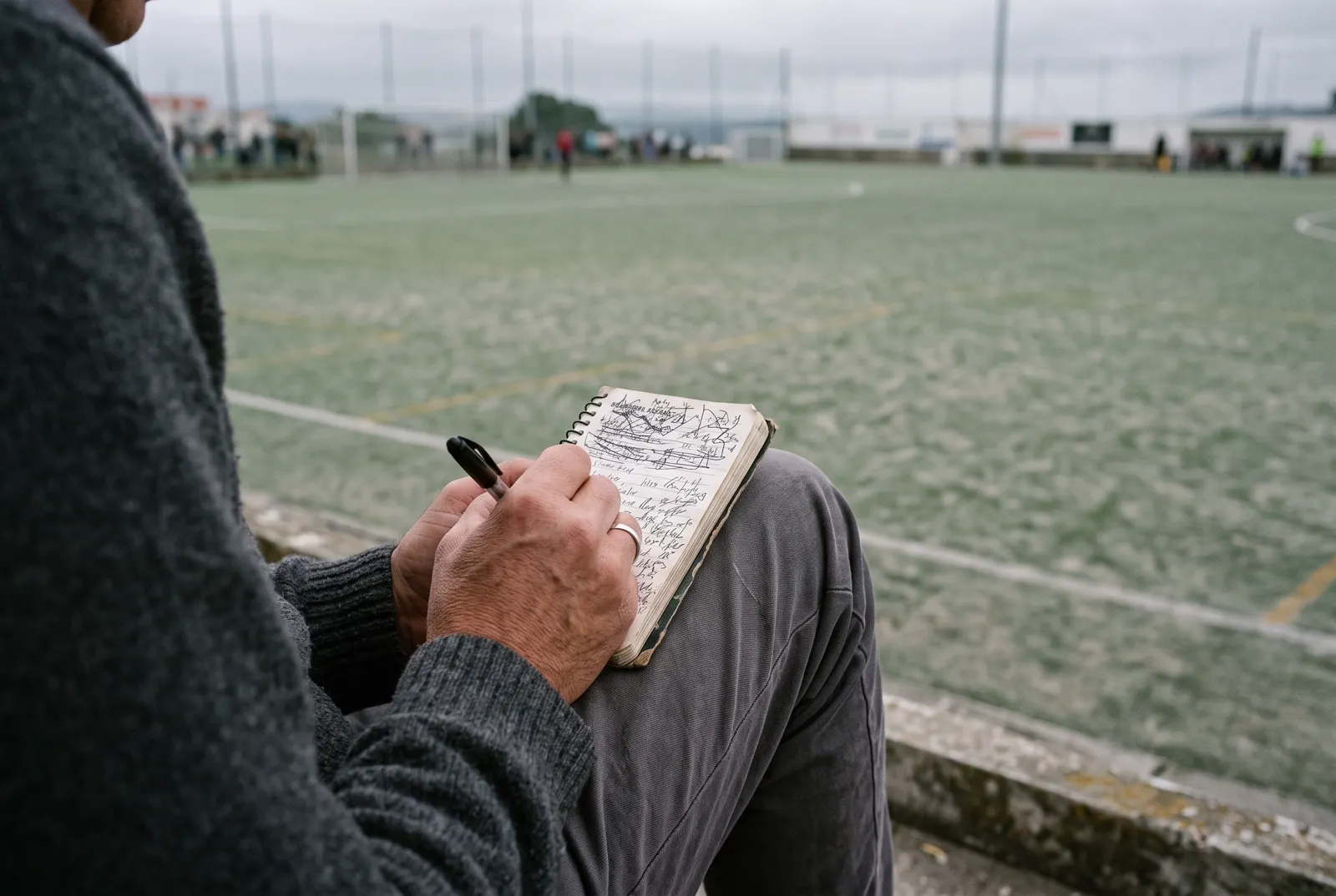 Mão a escrever num caderno pousado no joelho, à beira de um campo de relva sintética